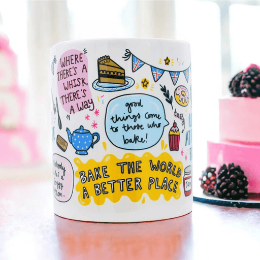 White ceramic mug featuring cheerful baking illustrations, including a slice of cake, teapot, and British bunting, with motivational quotes including "Where there's a whisk, there's a way" and "Bake the world a better place" shown on pink surface with berries in background.