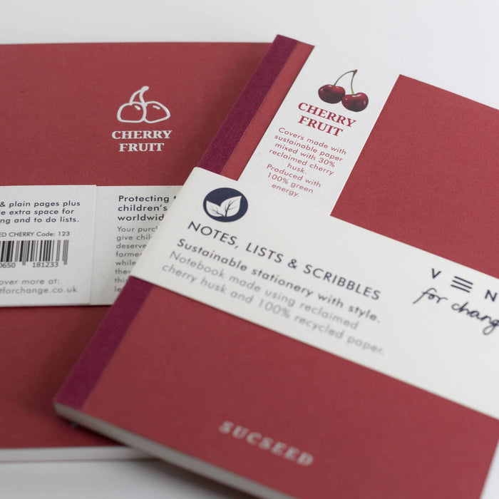 Close-up detail of two red cherry husk notebooks showing branding elements including the Cherry Fruit logo, product information band, and SUCSEED text. The barcode and VENT for change logo are visible on the back cover.