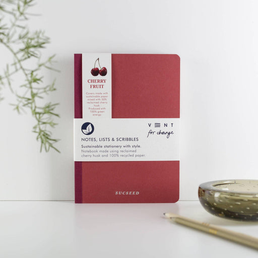 A red A5 recycled notebook with cherry fruit logo and 'SUCSEED' embossed in silver on the cover, shown against a white background with decorative green foliage. The notebook features a band displaying sustainable stationery details and the VENT for change branding.