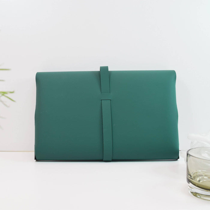 Emerald green recycled leather document wallet shown from side angle, displaying its sleek strap closure system. A glass with clear liquid visible at the edge of frame.