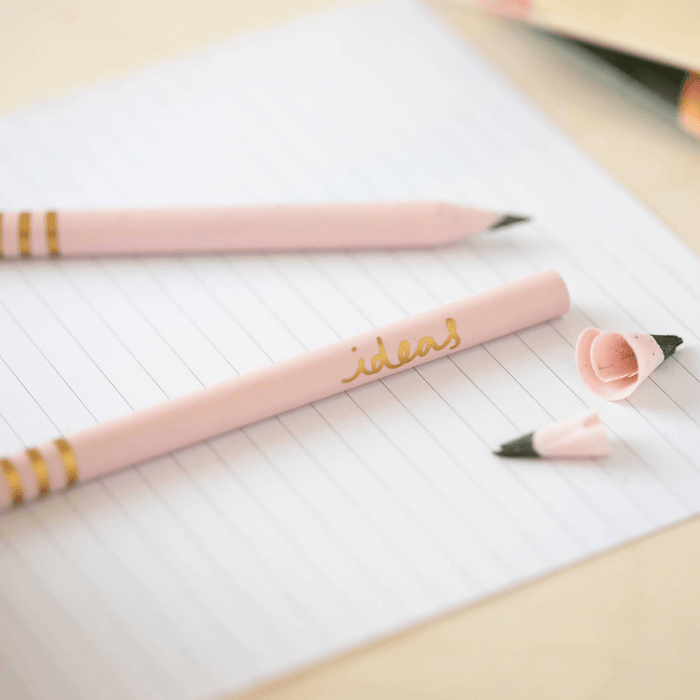 Close-up of pink recycled pencils with gold 'ideas' text and striped detailing on lined paper. Pencil shavings visible showing the unique recycled material construction.