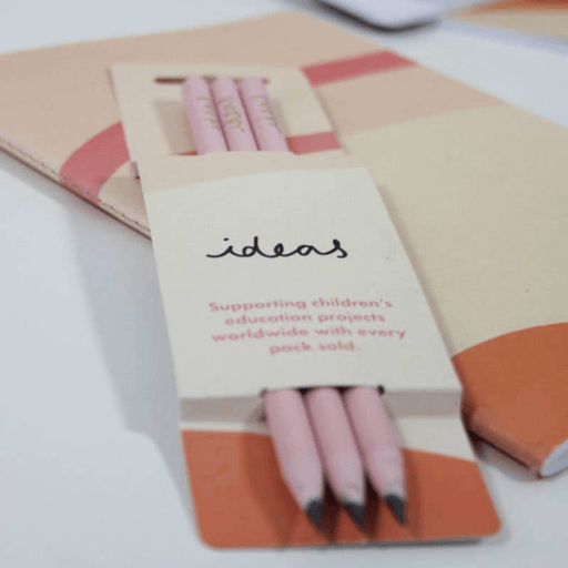 Three pink recycled pencils displayed in a cream-coloured sleeve with "ideas" handwritten text and coral accents. Small text mentions supporting children's education projects worldwide.