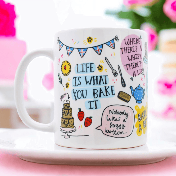 White ceramic mug with playful baking illustrations including British bunting, a tiered cake, strawberries, and teapot, featuring the quote "Life is what you bake it" and other baking puns, displayed on a pink surface with soft floral background.