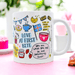 White ceramic mug featuring whimsical baking illustrations including British bunting, cookies, jam jar, strawberries, and playful text reading "Love at First Bite" and "Life is Short... Lick the Bowl!" displayed on pink surface with blurred floral background.