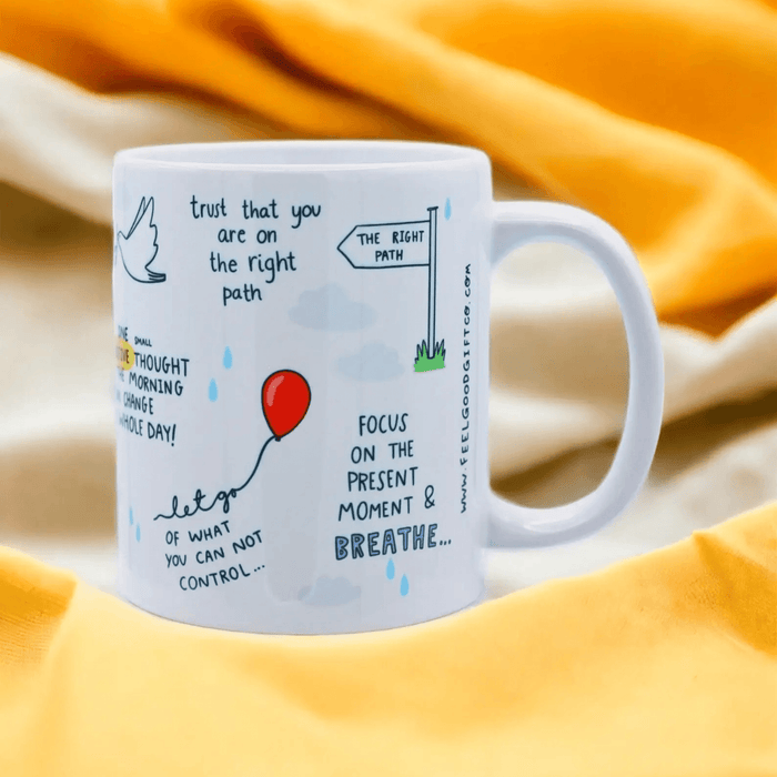 White ceramic mug featuring mindful affirmations including "trust that you are on the right path" and "focus on the present moment & breathe," decorated with a red balloon, signpost, and dove illustrations. Photographed on a warm yellow fabric background.