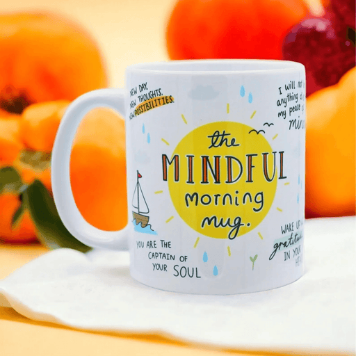 White ceramic mug with whimsical illustrations featuring "The Mindful Morning Mug" text in a yellow sun design, surrounded by positive affirmations including "You are the captain of your soul" with a small sailboat illustration. Photographed on a white surface with orange citrus fruits in the background.