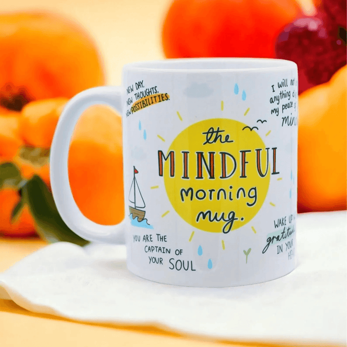 White ceramic mug with whimsical illustrations featuring "The Mindful Morning Mug" text in a yellow sun design, surrounded by positive affirmations including "You are the captain of your soul" with a small sailboat illustration. Photographed on a white surface with orange citrus fruits in the background.