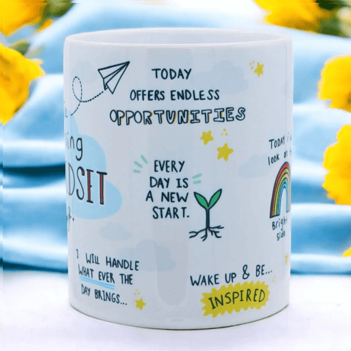 Close-up of white ceramic mug featuring hand-drawn inspirational quotes including "Today offers endless opportunities", "Every day is a new start", and "Wake up & be inspired", decorated with illustrations of a paper plane, rainbow, and sprouting plant, photographed against a blue background with sunflowers.