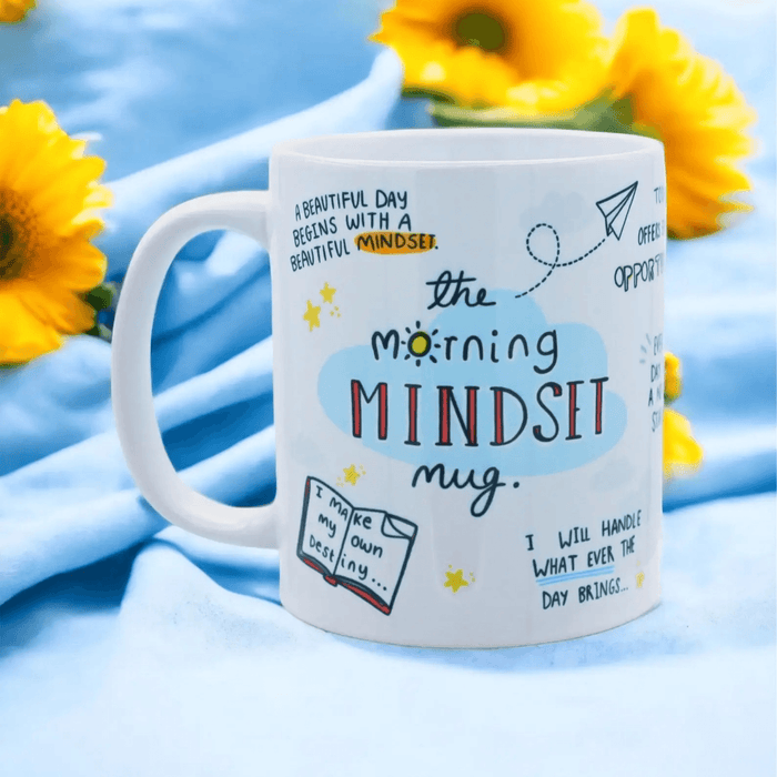 White ceramic mug with inspirational hand-drawn illustrations and text including "A beautiful day begins with a beautiful mindset" and "The Morning MINDSET mug", photographed against a blue background with bright yellow sunflowers.