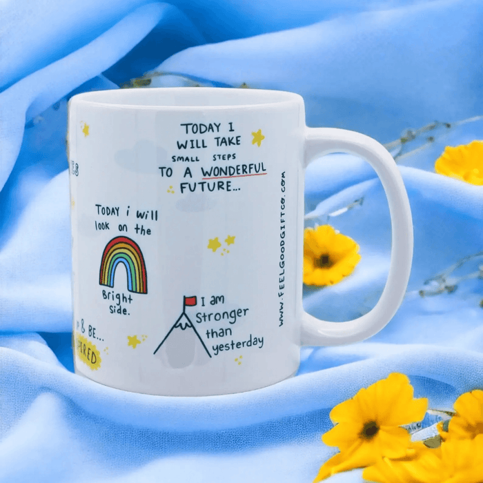 White ceramic mug with positive affirmations including "Today I will take small steps to a wonderful future" and "I am stronger than yesterday", featuring hand-drawn rainbow and mountain illustrations, styled with blue fabric and yellow flowers.