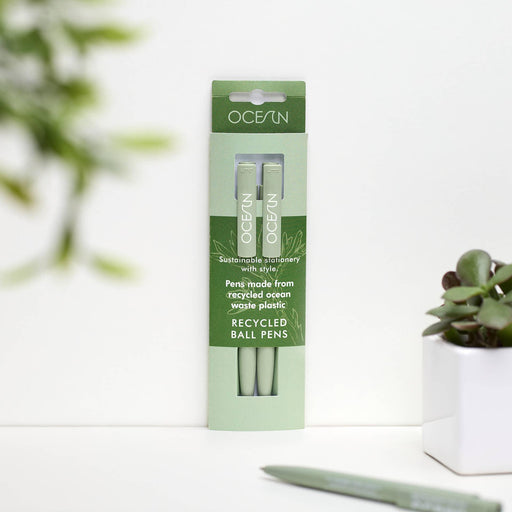 A pack of two mint green Ocean Collection recycled pens in eco-friendly packaging, displayed on a white surface with a succulent plant in the background. The packaging features green and white branding with text highlighting the pens are made from recycled ocean waste plastic.