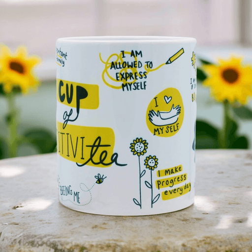 White ceramic mug with positive affirmations including "I am allowed to express myself" and "I make progress every day" in yellow and black hand-lettering, decorated with doodles of flowers and bees. Sunflowers visible in soft focus background.