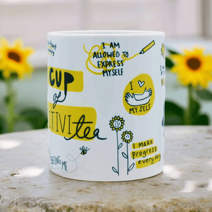 White ceramic mug with positive affirmations including "I am allowed to express myself" and "I make progress every day" in yellow and black hand-lettering, decorated with doodles of flowers and bees. Sunflowers visible in soft focus background.