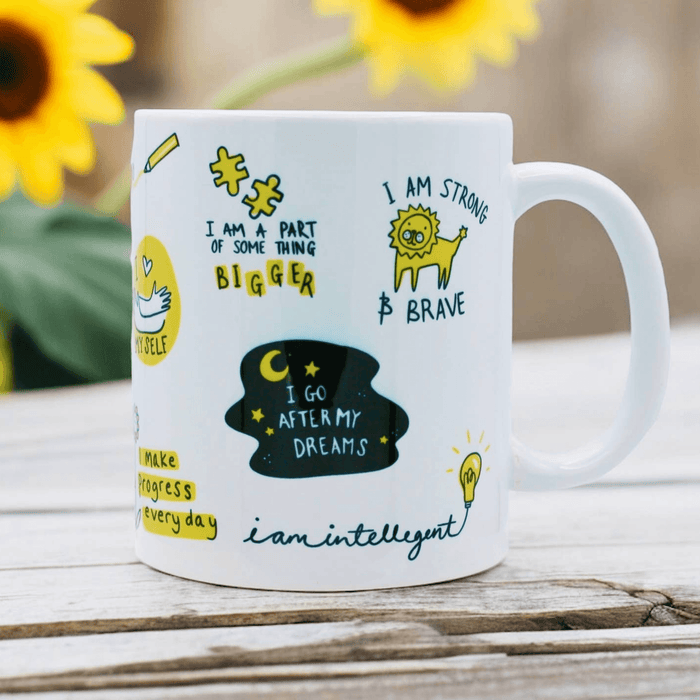 White ceramic mug featuring encouraging affirmations including "I am part of something bigger" and "I go after my dreams" with whimsical illustrations including a lion, light bulb, and stars. Photographed with sunflowers in background on wooden surface.