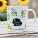 White ceramic mug featuring encouraging affirmations including "I am part of something bigger" and "I go after my dreams" with whimsical illustrations including a lion, light bulb, and stars. Photographed with sunflowers in background on wooden surface.