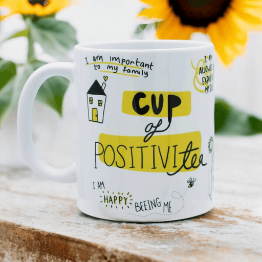 White ceramic mug with hand-drawn positive affirmations and illustrations, featuring "Cup of Positivitea" in yellow and black text, surrounded by happy quotes and a small house drawing. Sunflower visible in background.