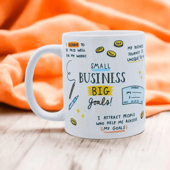 White ceramic mug with hand-drawn entrepreneurial affirmations and doodles including "Small Business BIG goals!" text, coins, and positive business mantras on an orange fabric background