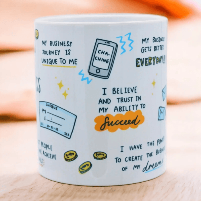 Close-up side view of white ceramic mug featuring motivational business affirmations, including "I believe and trust in my ability to succeed" and "My business gets better everyday" along with illustrations of a phone, coins, and cheque