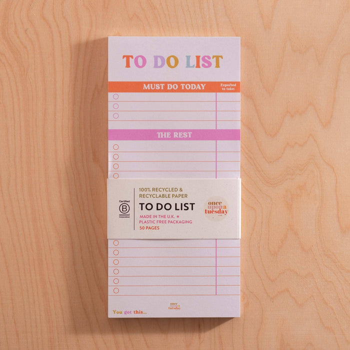 To Do List pad displayed on a wooden surface showing the belly band with product details including "100% Recycled & Recyclable Paper", "Made in the UK", and "Plastic Free Packaging". The pad's sections for "Must Do Today" and "The Rest" are visible with their orange and pink headers.