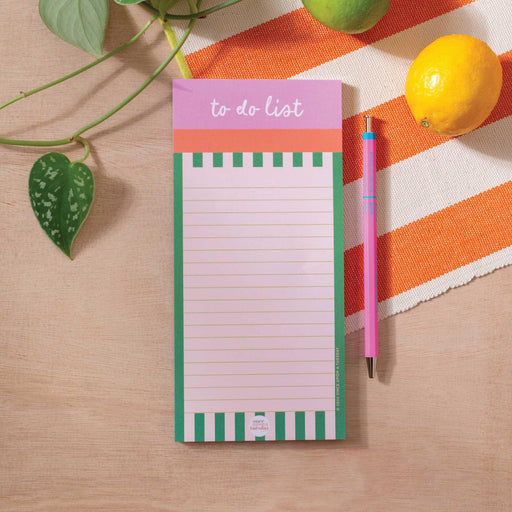 A Cabana Stripe To Do List Pad with green and white stripes and pink header, displayed on a wooden desk surface with an orange striped textile, pink pen, lemon, and potted plant leaf in view