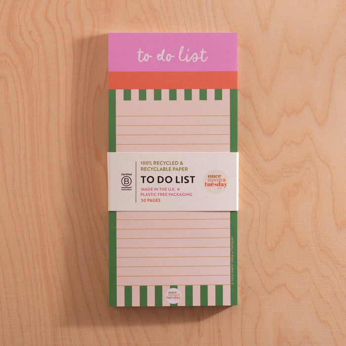 A Cabana Stripe To Do List Pad with pink and green design, shown with its belly band packaging that displays recycled paper certification and 'Made in the UK' text, photographed on a wooden surface