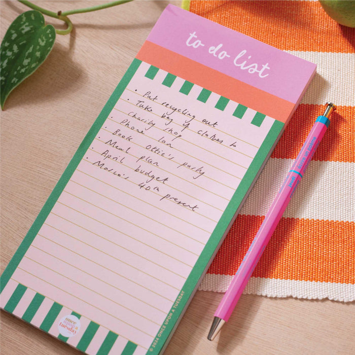 A filled-in Cabana Stripe To Do List Pad showing a handwritten task list, displayed with a pink and blue pen on a wooden surface with an orange striped cloth and plant leaf