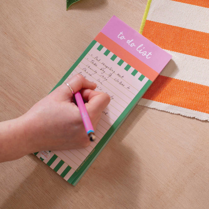 Hand writing on a green and white striped To Do List Pad with pink header, using a pink and blue pen. The pad is shown on a wooden desk with an orange striped cloth visible