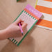 Hand writing on a green and white striped To Do List Pad with pink header, using a pink and blue pen. The pad is shown on a wooden desk with an orange striped cloth visible