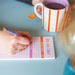 Person writing on the To Do List pad during a morning planning session, with a striped orange and white coffee mug in the background. The pad's colorful headers are visible as they plan their tasks for the day.