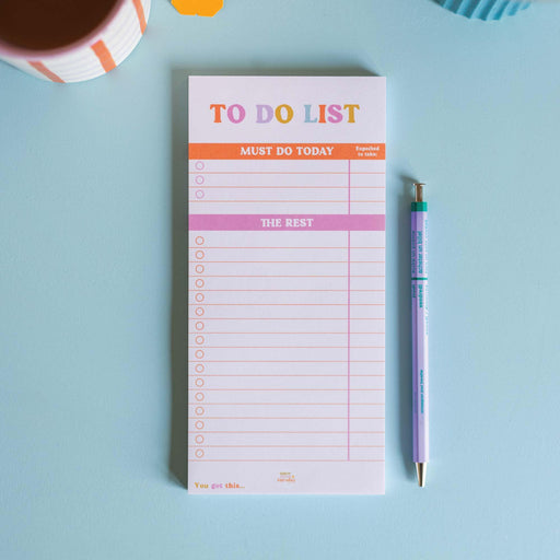 A minimalist To Do List pad with pink and orange text layout featuring "Must Do Today" and "The Rest" sections, displayed on a light blue desk surface next to a pastel mechanical pencil. The pad shows clean lined spaces for task writing.