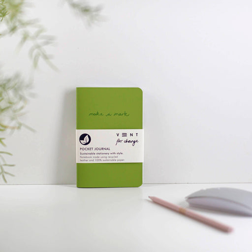 A bright green A6 journal with "make a mark" text on the cover, displayed against a white background with soft green foliage in the corner. A pencil rests beside the notebook. The journal features the VENT for change brand label.