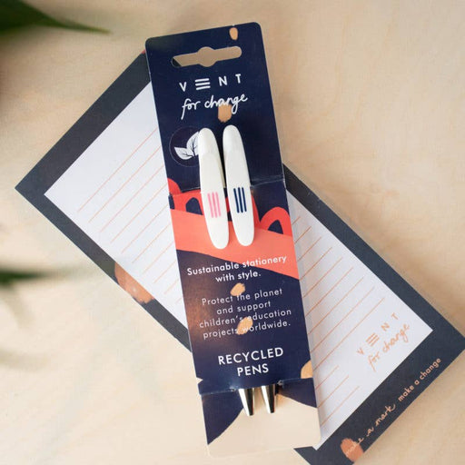 Two white recycled ball pens with black accents displayed on navy blue VENT For Change packaging card, shown alongside lined notepaper. The packaging features coral geometric designs and text highlighting sustainable stationery with style.