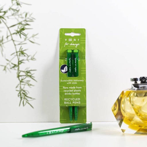 A pack of two green VENT For Change recycled ballpoint pens in retail packaging, displayed against a white background with decorative bamboo leaves and a yellow glass vase. One pen is removed from the packaging to show the sleek design.