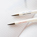 Two white VENT For Change pens with "make a change" text in blue and red lying on lined paper. The silver pen tips are shown in focus, demonstrating writing position.