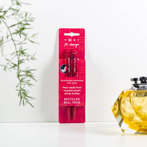 Two black ball pens in bright pink recycled packaging from VENT For Change, displayed against a light background with decorative foliage and amber glass bottle