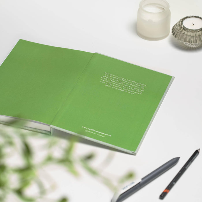 Open VENT notebook showing bright green interior pages with subtle text detail, displayed on white surface with pencils, glass tea light holders and soft foliage in frame.