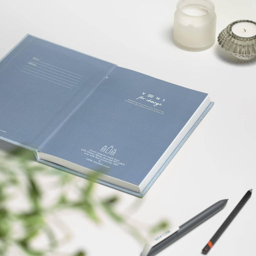 Open VENT Ocean Waste A5 Notebook showing blue-grey interior pages with minimalist branding, styled on white desk with pencils, tealight holders and greenery, modern workspace setting