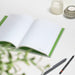 Opened VENT notebook showing crisp white lined pages with green binding edge, styled with pencils and candle holders, with blurred green foliage in foreground.