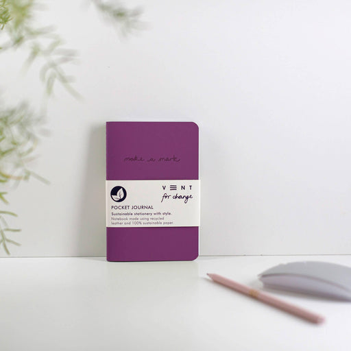 A purple A6 VENT pocket journal with recycled leather cover, featuring debossed 'make a mark' text and white paper band, displayed on a white surface with minimal styling including a pencil and leaves in soft focus