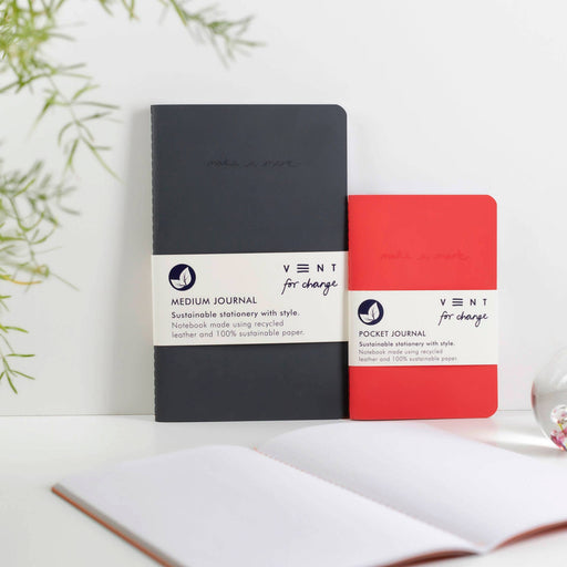 Two VENT recycled leather journals - one in charcoal black and one in coral red - displayed on a white surface alongside an open notebook. Green plant leaves visible in the background.