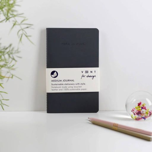 A slim charcoal-coloured recycled leather journal with VENT branding band sits on a white surface beside a pencil and decorative glass ornament. Green plant leaves visible in the background.