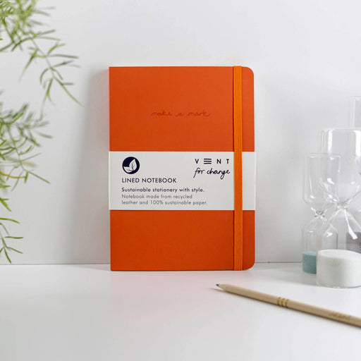 Orange recycled leather A5 notebook by VENT lying on a white desk with bamboo plant and pencil. Features "make a mark" debossing and elastic closure. Shown with product band highlighting sustainable materials and ethical production.