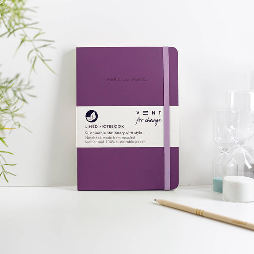 A purple A5 notebook with recycled leather cover lying on a white surface. The notebook features a light grey elastic closure band and "make a mark" embossed text. VENT's branded paper band shows "Lined Notebook" text. A wooden pencil and plant decoration visible in the scene.