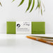 A green recycled leather pen pouch lying flat on a white surface with bamboo leaves visible in the background. The pouch features a white band with VENT for change branding and a leaf logo. Two wooden pencils are placed in the foreground, slightly out of focus.