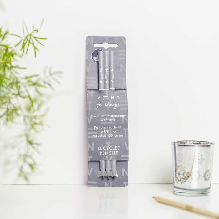 Pack of three dusty blue recycled pencils in VENT's grey packaging, displayed against a white background with decorative bamboo leaves and a glass tea light holder. The sustainable stationery packaging features the VENT logo and "for change" tagline.