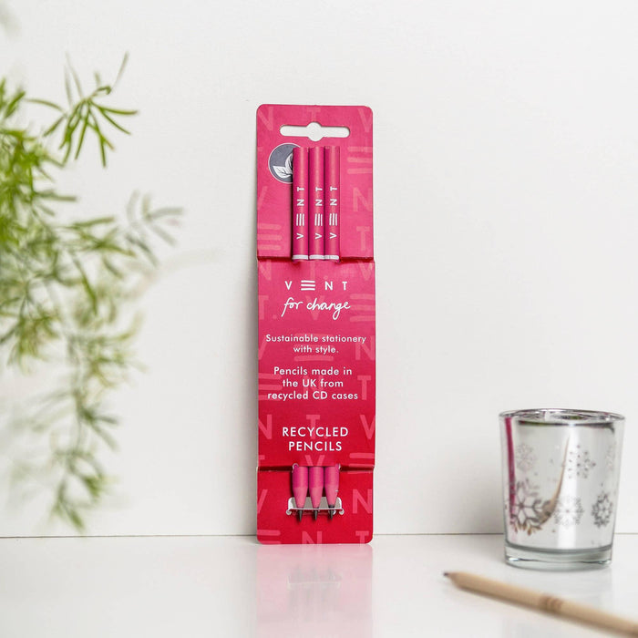 A pack of three bright pink VENT recycled pencils in pink packaging, displayed on a white desk surface next to a decorative glass candle holder and plant. The packaging features "for change" text and highlights that the pencils are made in the UK from recycled CD cases.