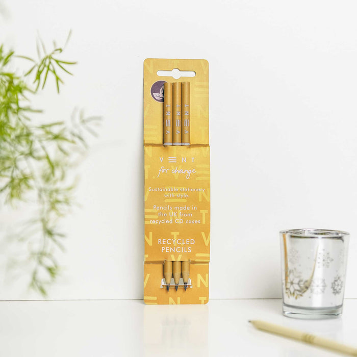 Pack of three yellow recycled pencils in VENT's signature packaging, displayed upright against a white background with decorative plant and glass in the scene. The yellow packaging features the VENT for change branding and recycled pencils text.