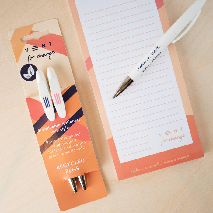 VENT sustainable pen packaging displayed alongside a coral-bordered notepad. The packaging shows two white recycled pens with the VENT "for change" branding and sustainability message. One pen is shown removed from the packaging and placed on the lined notepad, demonstrating the "make a mark make a change" text on its body.