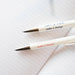 Close-up of two VENT recycled pens laid on lined paper, showing their "make a mark make a change" message in navy blue and coral pink text respectively. The silver chrome pen tips contrast against the crisp white notepad lines.