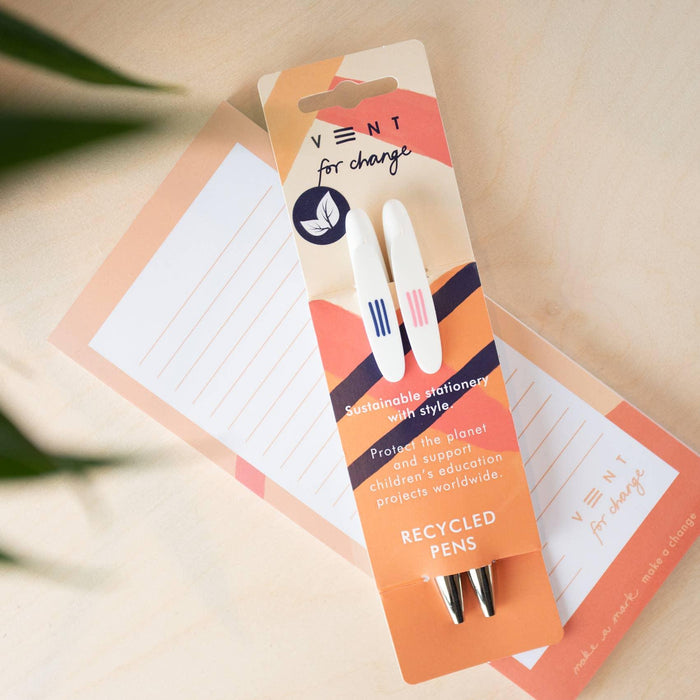 Two white recycled pens displayed on a coral-coloured VENT branded card packaging. The card features modern geometric shapes in coral, white and navy, with text about sustainable stationery and children's education projects. A lined notepad and plant leaf are partially visible in the background.